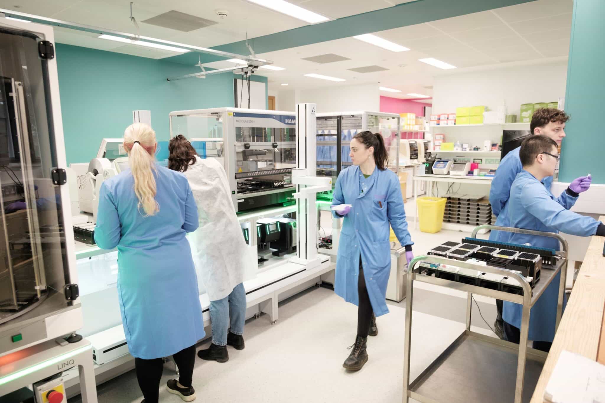 A group of people in a mixture of blue and white lab coats work around a robotic lab equipment.